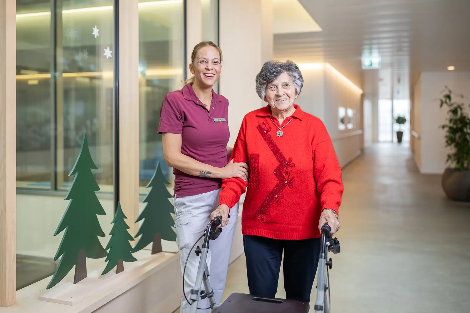 Eine Mitarbeitende unterstuetzt laechelnd eine Bewohnerin mit Rollator in einem hellen Gang.