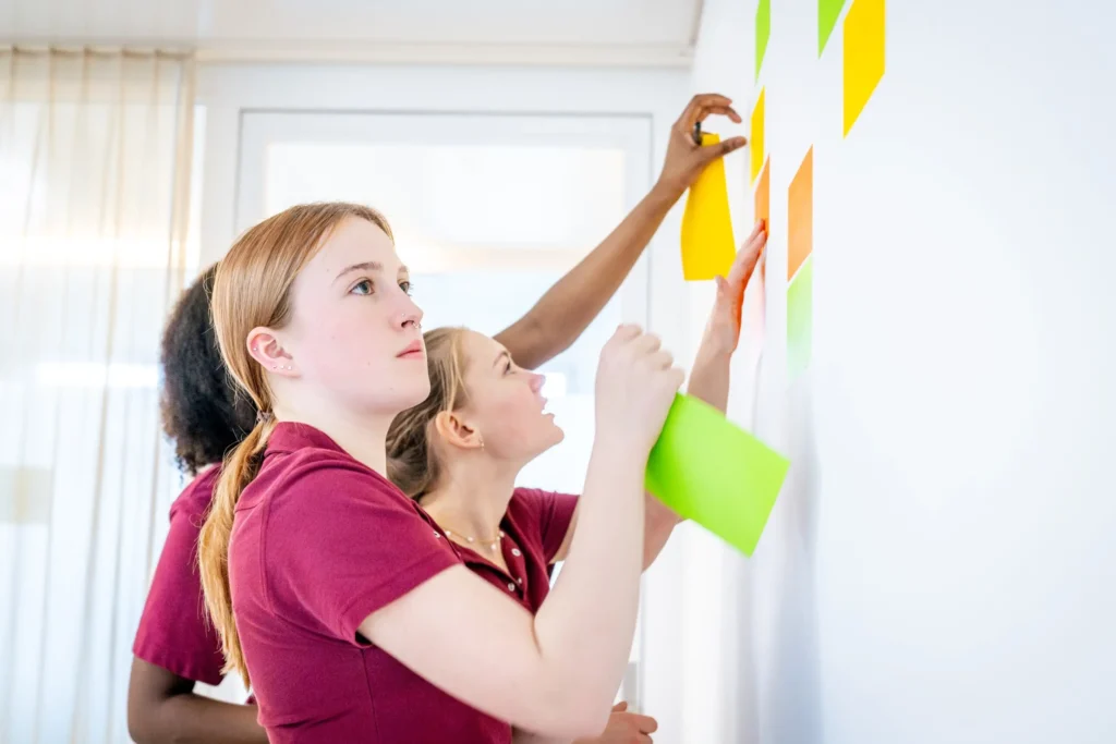Drei Auszubildende in der Pflege befestigen bunte Lernkarten an einer Wand und tauschen sich über Inhalte aus.