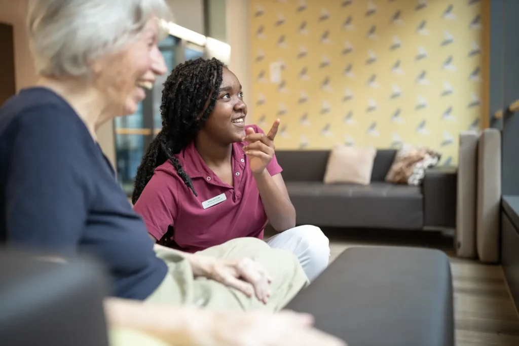 Eine lernende Fachperson Betreuung lacht mit einer Bewohnerin in einer gemütlichen Lounge.