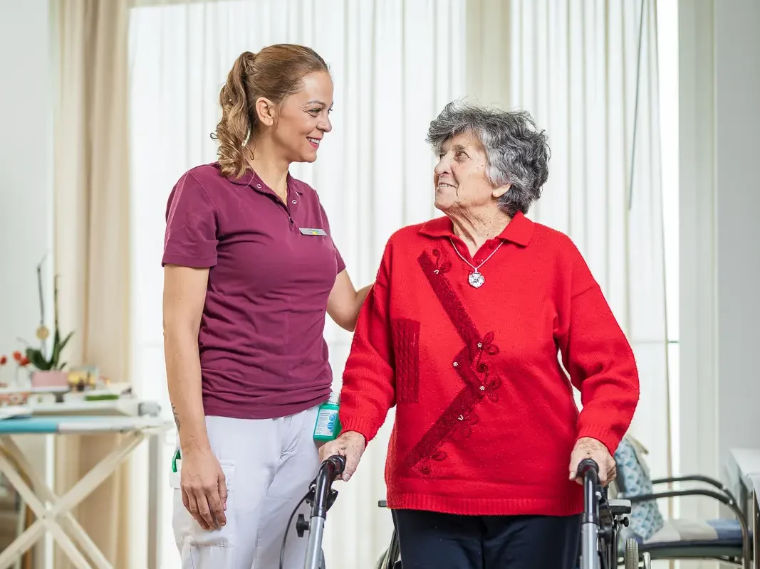 Eine Mitarbeitende begleitet eine Bewohnerin mit Rollator und schenkt ihr ein freundliches Laecheln.